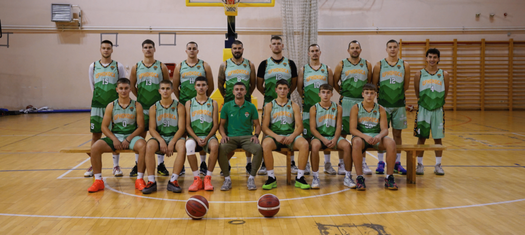 Tim košarkaša kk crnokosa u zelenim uniformama stoji i sedi u sportskoj sali. Dve lopte su ispred njih.