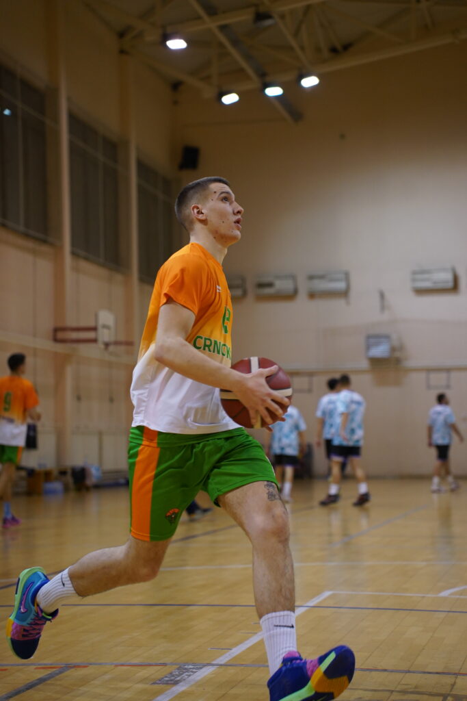 Košarkaš u narandžastoj i zelenoj opremi drži loptu na terenu.