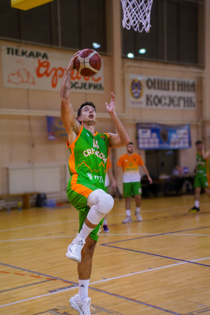 Košarkaš u zeleno-narandžastom dresu skače sa loptom prema košu.