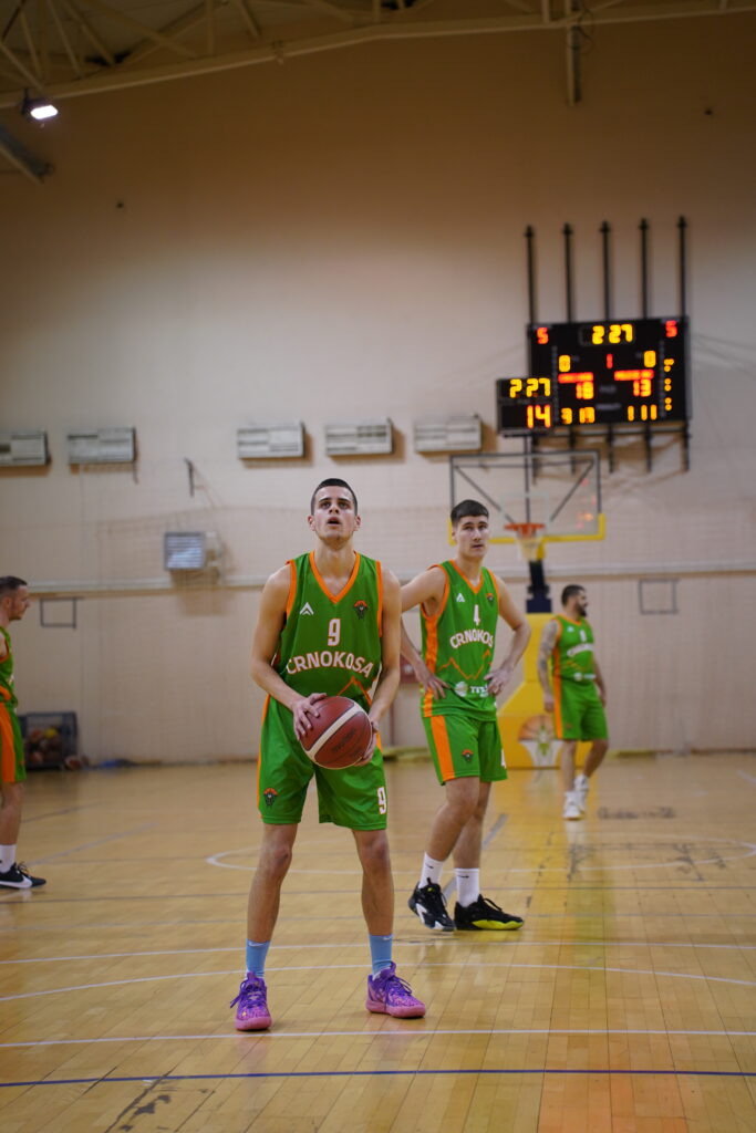 Košarkaš u zelenoj uniformi drži loptu, priprema se za šut.