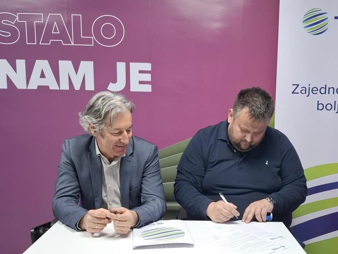 Dva muškarca potpisuju dokument za stolom, ljubičasta pozadina